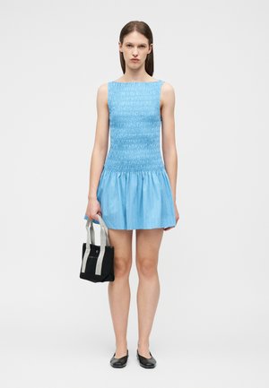 Nő ujjatlan, kék fodros miniruhában, fekete lapos cipőben, kezében kis fekete és szürke táskával, világos egyszínű háttér előtt áll.