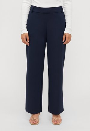 Femme portant un pantalon large taille haute bleu marine, des sandales beiges à brides et un haut blanc à manches longues.