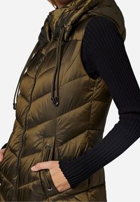 Oliwkowa pikowana kamizelka z kapturem, z zamkiem błyskawicznym z przodu, ozdobiona sznurkami oraz o diamentowym pikowaniu. Widoczne czarne, prążkowane rękawy.