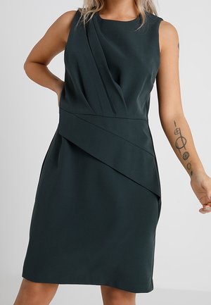Robe sans manches vert foncé avec un design superposé asymétrique au niveau de la taille et de la poitrine, portée par une personne avec des tatouages sur le bras droit.