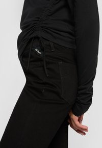 Person in schwarzem gerafftem Oberteil und schwarzen Jeans, bei denen das "REPLAY"-Label auf der Vorderseite der Tasche sichtbar ist, Hand entspannt an der Seite.