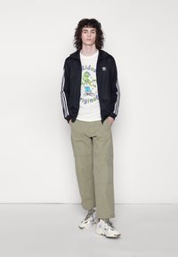 Svart Adidas zip-up jakke med hvite striper, grafisk hvit t-skjorte med en tegneserie, grønn bukse i cargo-stil og mønstrede joggesko.