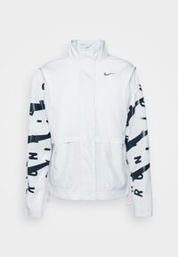 Veste blanche Nike avec col montant, fermeture éclair intégrale sur le devant, lettrage abstrait noir sur les manches et logo Nike sur la poitrine gauche.