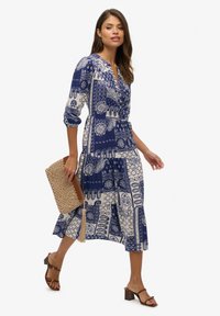 Navyblaues gemustertes Kleid mit Knopfdetails, Dreiviertelärmeln und einem gestuften Saum, kombiniert mit einer beigefarbenen geflochtenen Clutch und braunen Absatzschuhen.