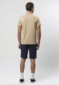 Beiges T-Shirt mit kurzen Ärmeln und rundem Ausschnitt, kombiniert mit marineblauen Shorts. Das Modell trägt weiße Socken und dunkle Turnschuhe.