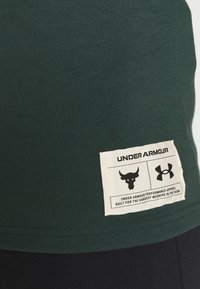 Camiseta de algodón verde con etiqueta beis, gráficos y texto en negro, dobladillo cosido y diseño ajustado.