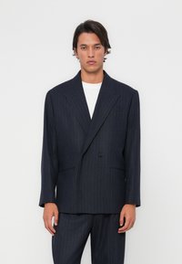 Blazer à rayures marine avec un design croisé, des revers crantés et des poches avant ; associé à un T-shirt blanc et un pantalon assorti.