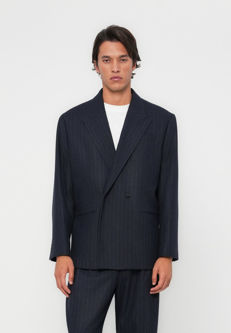 Blazer à rayures marine avec un design croisé, des revers crantés et des poches avant ; associé à un T-shirt blanc et un pantalon assorti.