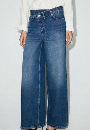 Persona con vaqueros azules de pierna ancha y camisa blanca de manga larga, de pie contra un fondo claro y liso.