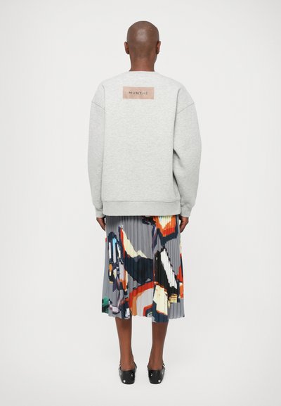 Sivá mikina s obdĺžnikovou nášivkou s nápisom "MUNTHE." V páre s farebnou plisovanou sukňou s abstraktnými vzormi a rôznymi farbami.