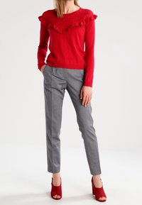 Rode trui met ruches aan de halslijn, gecombineerd met grijze, op maat gemaakte broek. Model draagt rode open schoenen met open teen. Neutrale achtergrond.