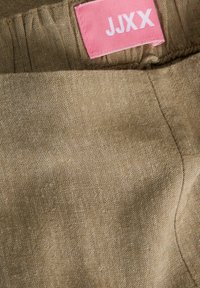 Μπεζ υφασμάτινη υφή με ραφές και μια ροζ ετικέτα ρούχων που δείχνει λευκά γράμματα "JJXX" ραμμένα στη μέση της ζώνης.