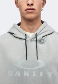 Νεαρό άτομο που φοράει ανοιχτό γκρι hoodie Oakley με μαύρα κορδόνια, εμφανίζοντας τα χείλη και το πηγούνι σε απλό φόντο.