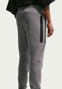 Makro af en person iført grå joggers med en sort lodret lynlåslomme på låret og et lille sort Nike-logo tæt på knæet.