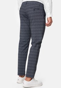 Mann trägt navy- und grau-karierte schlanke Hose, kombiniert mit weißen Sneakern und einem weißen Langarmshirt, seitlich stehend.