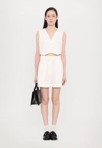 Top corto blanco con escote en V y botones en el frente, combinado con pantalones cortos blancos con pliegues y un bolso negro. La modelo lleva calcetines blancos y mocasines negros.