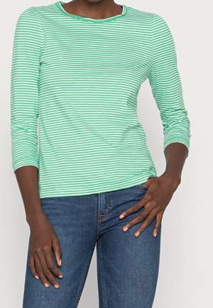 Vrouw met een groen-wit gestreept langärmelig shirt en een blauwe spijkerbroek, met de ene hand op haar heup en de andere langs haar zijde.