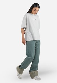 Lys grå oversized T-skjorte med korte ermer og en liten logo på brystet, kombinert med løstsittende grønne denimjeans og lyse sneakers.