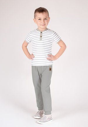 Biały t-shirt w paski z zielonym kołnierzem i guzikami, w połączeniu z szarymi dresami, na których znajduje się logo zwierzęcia, oraz wsuwane buty w kratkę.