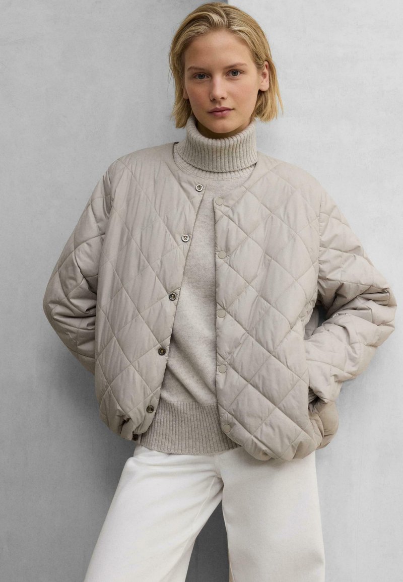 Beige gesteppte Bomberjacke mit Druckknöpfen über einem hellen Pullover. Sie verfügt über einen hohen gerippten Kragen und eine lockere Passform. Glatte Textur und weiches Material.