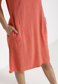 Korallklänning i lätt tyg, med två sidfickor och en avslappnad passform. Sömmen är rundad och texturen ser mjuk ut.