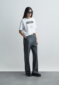 Balts T-krekls ar melnu "Moschino Milano" drukājumu, apvienots ar pelēkiem augstām biksēm un melnām kurpēm, papildināts ar tumšām saulesbrillēm.