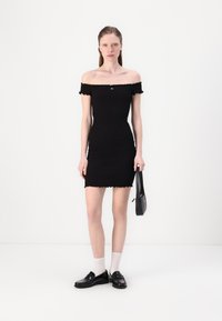 Robe noire décolletée, au design ajusté, en tissu texturé, avec des bords en forme de vagues. Le mannequin porte des mocassins et tient un petit sac noir.