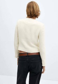 Veste courte texturée crème à manches longues, ourlet effiloché, portée sur un jean noir avec une ceinture marron, vue de dos.