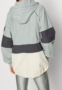 Chaqueta ligera con capucha en azul pálido, con paneles en gris oscuro y crema. Presenta puños elásticos y el logo impreso en la parte posterior.