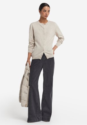 Beige stickad cardigan med rund halsringning, knappstängning och ribbade manschetter, i kombination med utsvängda mörka denimbyxor.