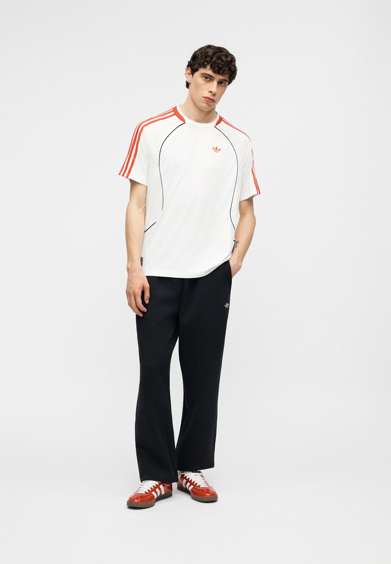 Junger Mann trägt ein weißes T-Shirt mit orangen Streifen, schwarze Hose und rote Adidas-Sneaker und steht vor einem schlichten Hintergrund.