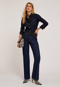 Donkere spijkerblouse met knoopsluiting aan de voorkant en borstzakken, gecombineerd met wijde donkere spijkerbroeken. Versterkt met een handtas met slangenprints en hakken.