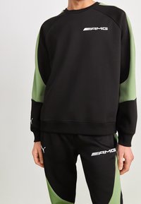 Osoba ubrana w czarną i zieloną bluzę z logo AMG oraz pasujące spodnie z logo Pumy, stojąca na jednolitym jasnym tle.