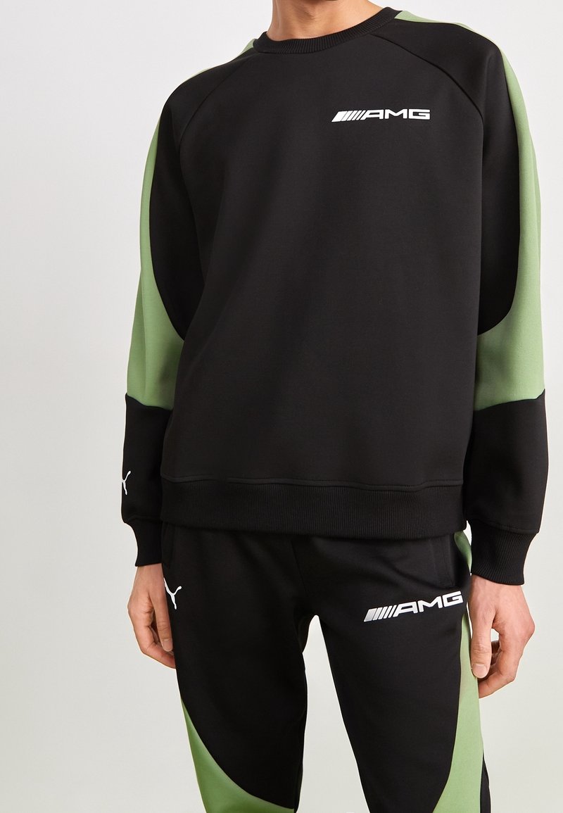 Osoba ubrana w czarną i zieloną bluzę z logo AMG oraz pasujące spodnie z logo Pumy, stojąca na jednolitym jasnym tle.