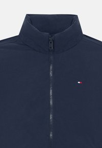 Veste zippée bleu marine avec un col haut, confectionnée dans un tissu lisse, arborant un petit logo rouge, blanc et bleu sur la poitrine gauche.