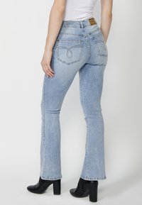 Jeans de mezclilla de corte acampanado en azul claro, con dos bolsillos traseros con costuras decorativas y un parche de cuero marrón en la cintura.