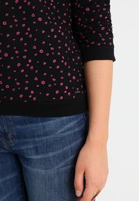 Felpa nera con motivo floreale rosa, polsini e orlo a coste. Indossata con jeans blu, mostrando un primo piano del maniche e della mano.