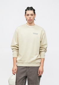 Μπεζ φούτερ με ριμπ μανσέτες και τελείωμα, με λογότυπο "PENFIELD" στο στήθος. Το ύφασμα φαίνεται μαλακό και με άνετη εφαρμογή.