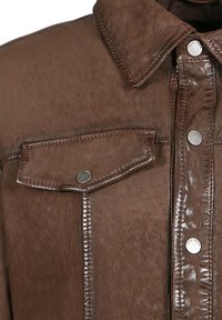 Detalje af brun læderjakke, der viser krave, knaplukket klaplomme og sølvfarvede trykknapper på frontstolpen.