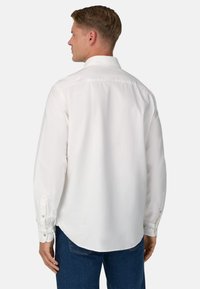 Uomo con i capelli corti che indossa una camicia bianca a maniche lunghe con bottoni e jeans blu, mostrato di spalle su uno sfondo neutro.