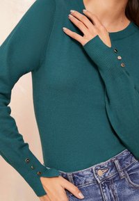 Pulover tricotat teal cu manșete striate și trei accente din butoni aurii. Design ajustat, textură moale, purtat cu pantaloni scurți din denim cu talie înaltă.