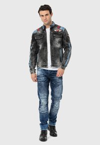Denimjack met vervaagde zwarte wassing, blauwe accenten, twee borstzakken en geborduurde details; gecombineerd met blauwe jeans en zwarte laarzen.