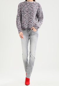 Žena oblečená v pestrobarevném pleteném svetru, světlých šedých džínách a červených kotníkových botách, stojící před jednoduchým světlým pozadím.