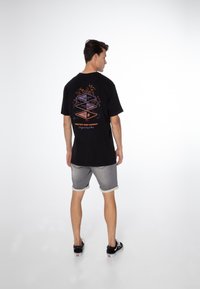 Zwart T-shirt met een kleurrijke grafische print op de achterkant, ontworpen met korte mouwen en een losse pasvorm. Draagt lichtgrijze shorts.