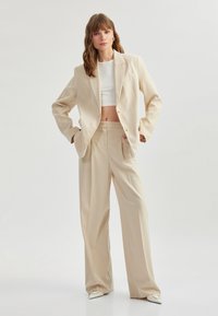Blazer beige su misura con bottoni dorati, abbinato a pantaloni a gamba larga a vita alta dello stesso colore, accompagnato da un crop top bianco e scarpe a punta.