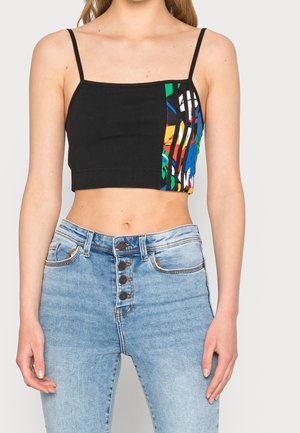 Kobieta w czarnym, krótkim topie na ramiączkach z kolorowym, abstrakcyjnym wzorem z jednej strony oraz w jasnoniebieskich, wysokim stanem i z przodem zapinanym na guziki jeansach.