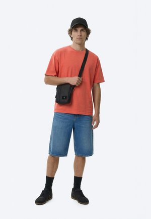 Fiatal férfi áll, fekete sapkát, narancssárga pólót, kék farmer rövidnadrágot, fekete tornacipőt, fekete zoknit és fekete kereszt testtáskát visel.