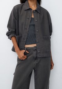 Giacca di denim grigia con chiusura a bottoni, due tasche frontali e maniche arrotolate, abbinata a un crop top a righe grigie e nere e pantaloni coordinati.
