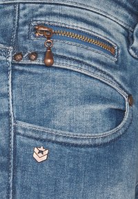 Modré džínové kalhoty s měděným zipem na kapse, měděnými kovovými doplňky a malou kovovou značkou na přední straně.