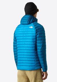 Blå isolerad jacka med huva, med ett quiltat mönster och en liten vit logotyp på ryggen. Slät textur och lätt material.